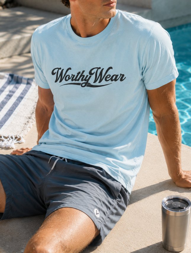 Light blue Worthy Wear basic tee with black logo, worn by a man sitting by a pool, paired with gray shorts.