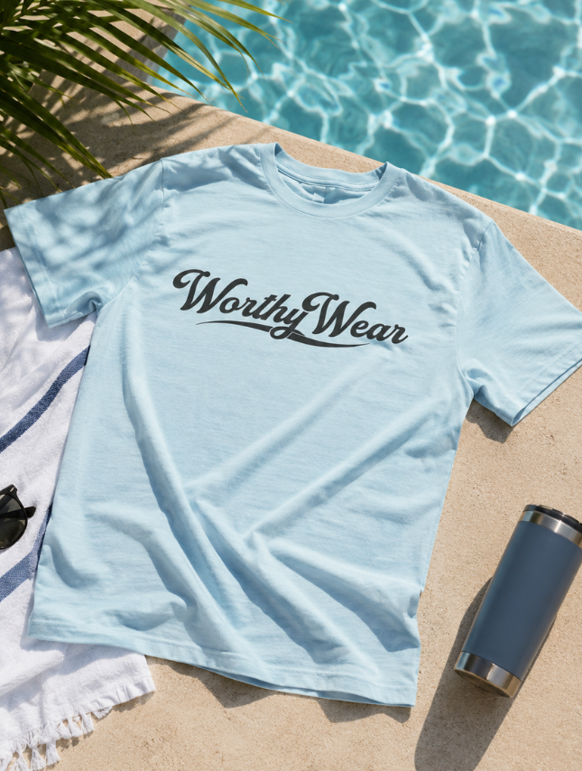 Light blue Worthy Wear basic tee with a black logo, placed on a tan surface near a pool and a towel.