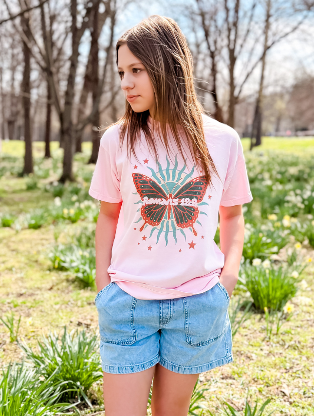 Youth tee in light pink featuring a butterfly graphic with "Romans 12:2" text, worn with denim shorts in a grassy outdoor ...