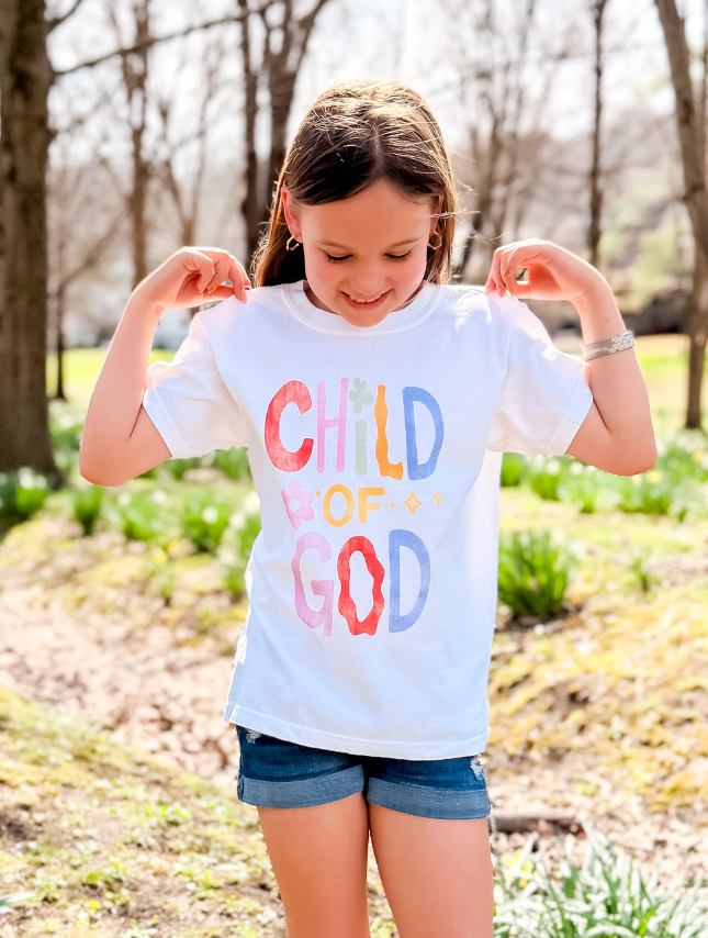 Youth tee featuring colorful "Child of God" text, worn by a girl outdoors in a sunny park setting.