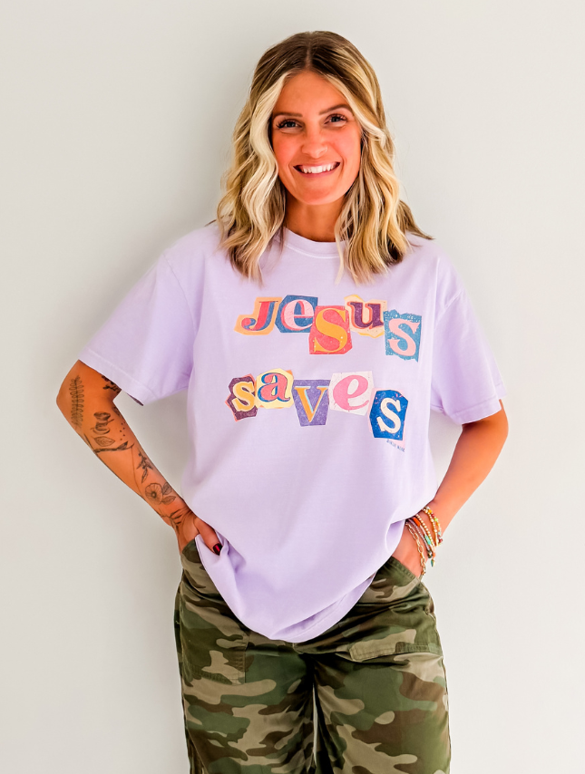 Jesus saves poster tee in lavender with colorful text, worn by a smiling woman in camo pants against a neutral background.