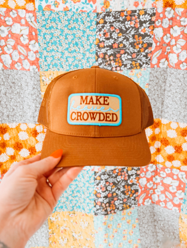 Brown cap with 'MAKE CROWDED' patch held against a colorful patchwork background
