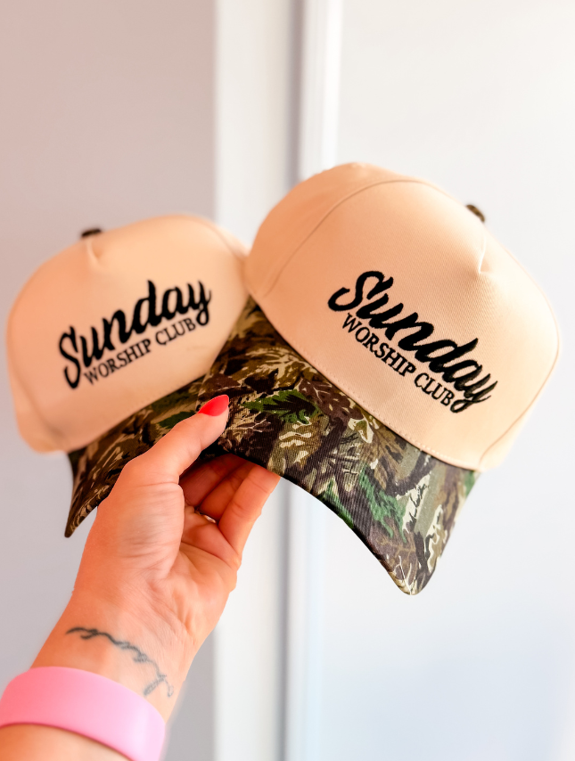Cream-colored Sunday Worship Club hat with a camouflage brim, held in a hand against a light background.
