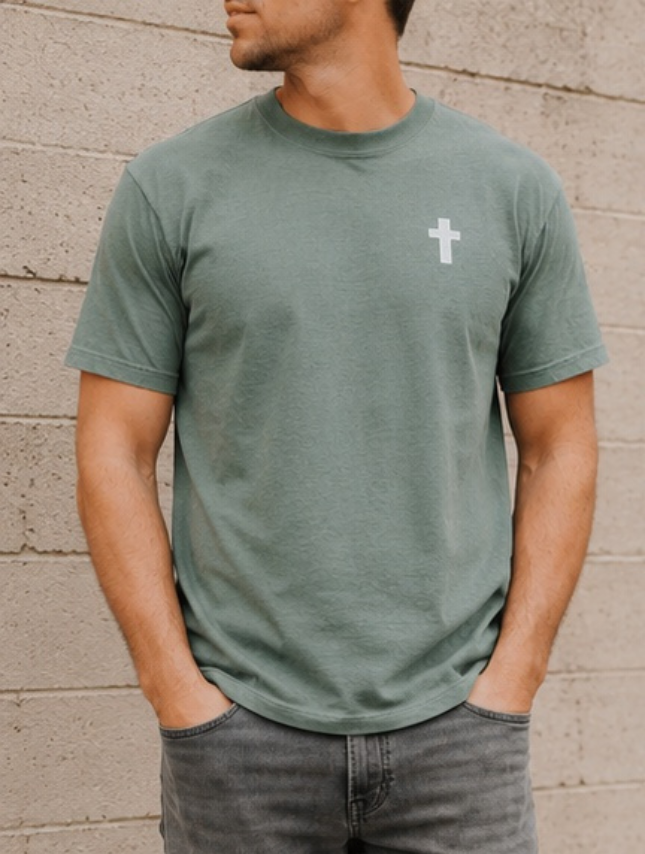 He Moves Mountains tee in moss green with a white cross on the left chest, worn by a man against a gray wall.